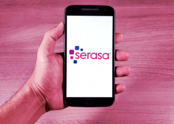 Mão de uma pessoa segurando um smartphone que exibe o logotipo colorido da Serasa em uma tela branca, com fundo de madeira clara em tons rosados.