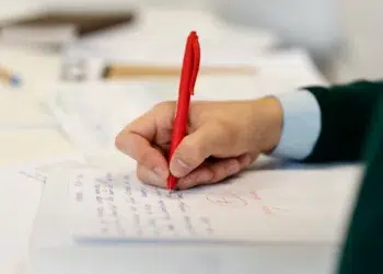 Mão segurando caneta vermelha escrevendo texto em folha branca