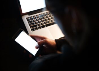 Silhueta de uma pessoa em ambiente escuro manuseando um smartphone com tela brilhante em frente a um notebook aberto, simbolizando o risco de crimes cibernéticos e roubo de dados pessoais.