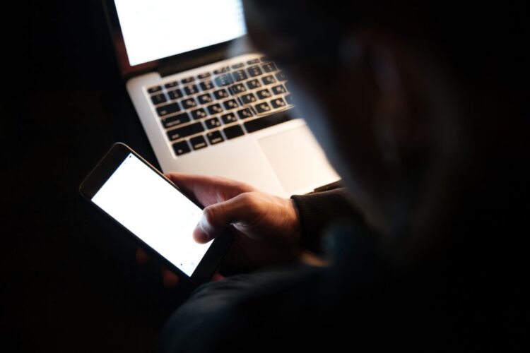 Golpistas usam seu nome e CPF para fazer cobranças falsas da Receita; saiba como se proteger 1 Silhueta de uma pessoa em ambiente escuro manuseando um smartphone com tela brilhante em frente a um notebook aberto, simbolizando o risco de crimes cibernéticos e roubo de dados pessoais.