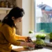 Mulher cuidando de temperos em uma horta dentro de apartamento, podando ervas em uma janela iluminada.