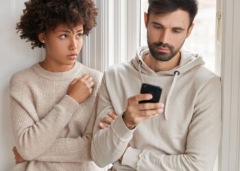 Um homem de barba e moletom cinza olha fixamente para o celular enquanto ignora uma mulher ao seu lado. A mulher, com cabelo cacheado volumoso e blusa de tricô bege, observa-o com uma expressão de dúvida e descontentamento. Ambos estão encostados em uma parede branca próxima a uma janela.