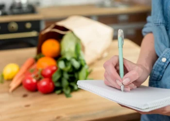 Pessoa anotando lista de compras em um caderno na cozinha com alimentos frescos sobre a mesa.