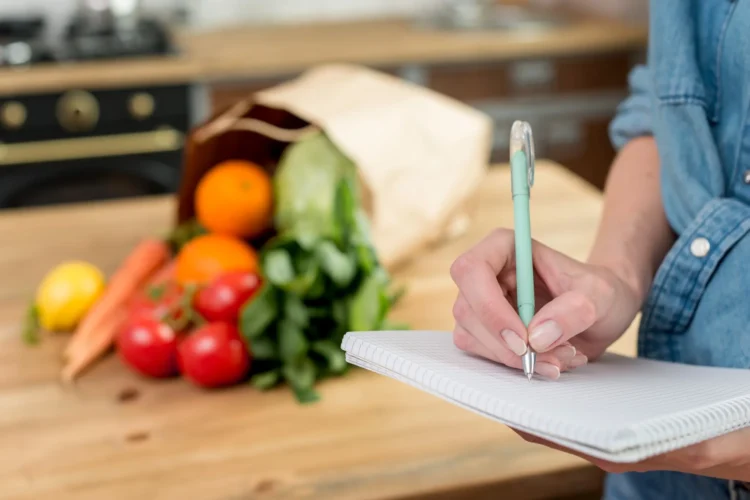 Pessoa anotando lista de compras em um caderno na cozinha com alimentos frescos sobre a mesa.