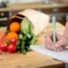 Pessoa anotando lista de compras em um caderno na cozinha com alimentos frescos sobre a mesa.