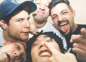 Foto de plano médio superior, em ângulo de selfie aproximada e descontraída, mostrando os rostos de quatro homens brancos e jovens. O homem no canto superior esquerdo usa um boné de beisebol azul-marinho. O homem no canto superior direito está sorrindo de forma ampla, com a língua para fora e o dedo indicador apontado para a câmera. O homem no canto inferior direito está fazendo uma careta engraçada, com o dedo indicador esquerdo no nariz. Apenas a parte superior da cabeça e o rosto do quarto homem são visíveis no canto inferior esquerdo. A iluminação é natural e o fundo é indistinto, em tom bege-claro.