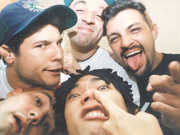 Esse detalhe surpreendente na exumação dos Mamonas Assassinas vai te emocionar! 1 Foto de plano médio superior, em ângulo de selfie aproximada e descontraída, mostrando os rostos de quatro homens brancos e jovens. O homem no canto superior esquerdo usa um boné de beisebol azul-marinho. O homem no canto superior direito está sorrindo de forma ampla, com a língua para fora e o dedo indicador apontado para a câmera. O homem no canto inferior direito está fazendo uma careta engraçada, com o dedo indicador esquerdo no nariz. Apenas a parte superior da cabeça e o rosto do quarto homem são visíveis no canto inferior esquerdo. A iluminação é natural e o fundo é indistinto, em tom bege-claro.