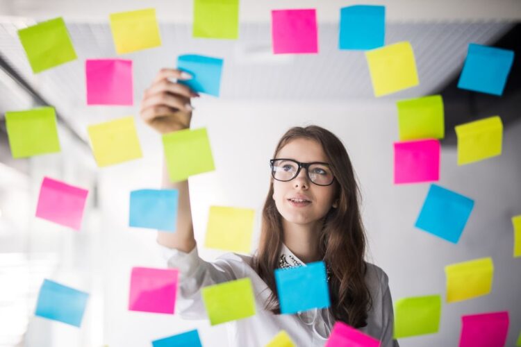 Mania de organização ou TOC? Entenda a diferença real segundo especialistas 1 Uma mulher jovem de óculos e camiseta preta organiza meticulosamente notas adesivas coloridas em um painel de vidro transparente. Ao fundo, um ambiente de escritório iluminado.