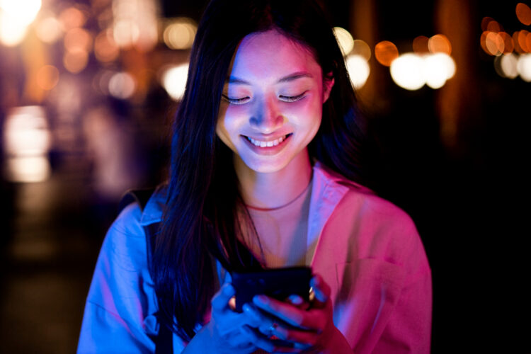 Mulher jovem sorrindo ao enviar mensagem no celular durante a noite com luzes desfocadas ao fundo