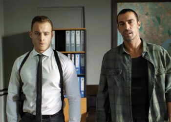 Dois homens adultos em um escritório com estantes de arquivos azuis ao fundo. O homem à esquerda, Kerem Bürsin, usa camisa social branca, gravata preta fina e um coldre de ombro preto. O homem à direita veste uma camisa de flanela xadrez verde sobre uma camiseta preta. Há um mapa-múndi na parede à direita.