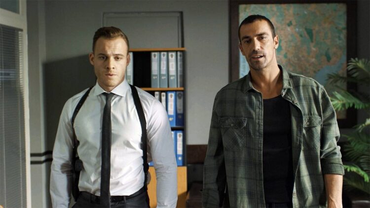 Dois homens adultos em um escritório com estantes de arquivos azuis ao fundo. O homem à esquerda, Kerem Bürsin, usa camisa social branca, gravata preta fina e um coldre de ombro preto. O homem à direita veste uma camisa de flanela xadrez verde sobre uma camiseta preta. Há um mapa-múndi na parede à direita.