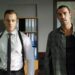 Dois homens adultos em um escritório com estantes de arquivos azuis ao fundo. O homem à esquerda, Kerem Bürsin, usa camisa social branca, gravata preta fina e um coldre de ombro preto. O homem à direita veste uma camisa de flanela xadrez verde sobre uma camiseta preta. Há um mapa-múndi na parede à direita.