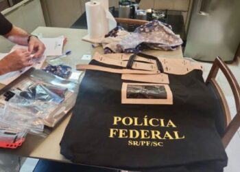 Policial Federal fardado analisando documentos sobre uma mesa preta com envelopes de evidências e uma maleta tática preta com a inscrição "POLÍCIA FEDERAL SR/PF/SC".
