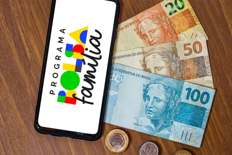 Bolsa Família de março 2026 começa dia 18 com novo calendário de pagamentos por NIS 1 Smartphone exibindo o logotipo colorido do Programa Bolsa Família ao lado de cédulas de 20, 50 e 100 reais e moedas sobre uma mesa de madeira.