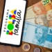 Smartphone exibindo o logotipo colorido do Programa Bolsa Família ao lado de cédulas de 20, 50 e 100 reais e moedas sobre uma mesa de madeira.