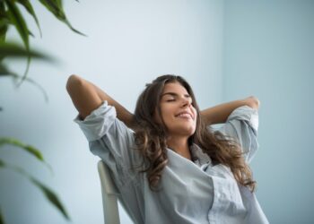 Mulher jovem de cabelos castanhos sentada em uma cadeira branca, com os olhos fechados e mãos atrás da cabeça em sinal de relaxamento, sorrindo levemente em um ambiente iluminado.