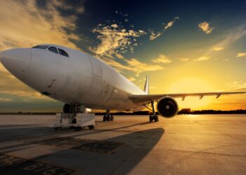 Visão em ângulo baixo de um grande avião comercial totalmente pintado de branco, estacionado na pista de um aeroporto com o sol se pondo ao fundo, criando silhuetas e tons alaranjados no céu.