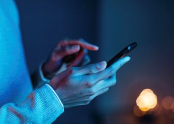 Close-up das mãos de uma pessoa segurando um smartphone em ambiente escuro com iluminação azul e ciano, enquanto interage com a tela sensível ao toque.