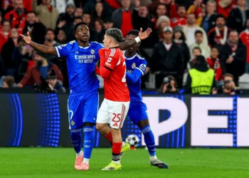 Atleta do Real Madrid levanta os braços em campo durante partida intensa, cercado por adversário e torcida atenta