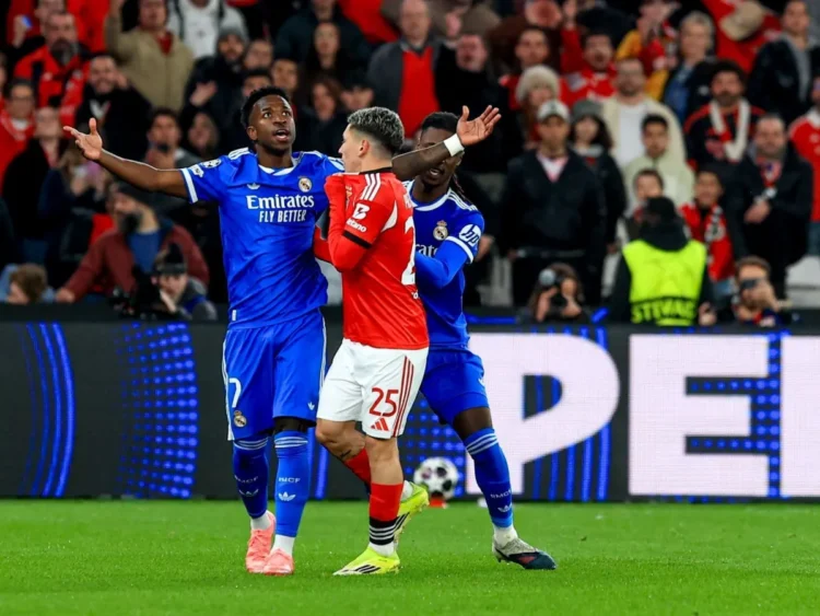 Racismo contra Vini Jr: Real Madrid envia provas à Uefa; veja nota 1 Atleta do Real Madrid levanta os braços em campo durante partida intensa, cercado por adversário e torcida atenta