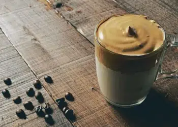Copo de café cremoso com camada de espuma dourada e grãos de café ao redor, servido sobre uma mesa de madeira.