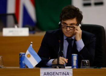 O presidente da Argentina, Javier Milei, sentado à mesa de uma conferência internacional. Ele veste terno escuro, camisa azul e gravata, ajustando seus óculos com a mão direita enquanto olha atentamente para frente. À sua frente, há uma pequena bandeira da Argentina, latas de bebida e uma placa com o nome do país.