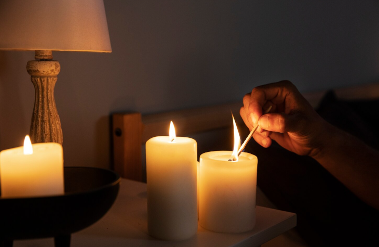 4 simpatias poderosas para atrair mais dinheiro neste mês de março 2 Foto em ambiente de baixa luminosidade focada em uma mão acendendo velas brancas cilíndricas sobre um móvel de madeira. Há três velas acesas que emanam uma luz amarelada e quente, criando uma atmosfera de paz e rito. Ao fundo, vê-se a base de um abajur clássico e parte da cabeceira de uma cama, sugerindo um ritual doméstico noturno.
