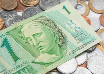 Close-up de uma cédula de um real verde e diversas moedas de Real de diferentes valores espalhadas sobre uma superfície branca. O foco principal está na efígie da República na nota, simbolizando a circulação de dinheiro e a riqueza.