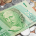 Close-up de uma cédula de um real verde e diversas moedas de Real de diferentes valores espalhadas sobre uma superfície branca. O foco principal está na efígie da República na nota, simbolizando a circulação de dinheiro e a riqueza.
