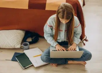 Jovem escrevendo em um laptop, com cadernos e xícara de café ao lado, em um ambiente de estudo.