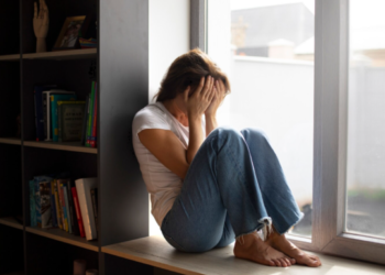 Uma mulher jovem, sentada de perfil no parapeito de uma janela interna, veste camiseta branca e calça jeans. Ela está com o corpo encolhido e cobre o rosto com as duas mãos em um gesto de angústia ou exaustão emocional. Ao lado, uma estante com livros reforça o ambiente doméstico de solidão.