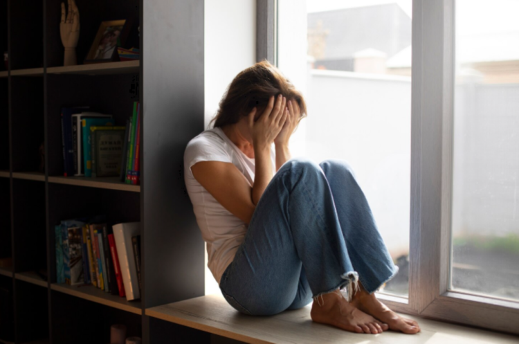 O "Efeito Domingo": Por que a ansiedade bate antes da semana começar e como evitá-la hoje 1 Uma mulher jovem, sentada de perfil no parapeito de uma janela interna, veste camiseta branca e calça jeans. Ela está com o corpo encolhido e cobre o rosto com as duas mãos em um gesto de angústia ou exaustão emocional. Ao lado, uma estante com livros reforça o ambiente doméstico de solidão.