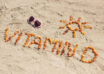 Palavra “Vitamina D” escrita na areia da praia com pequenas pedras alaranjadas, ao lado de um desenho de sol e óculos de sol em formato de coração.
