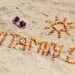 Palavra “Vitamina D” escrita na areia da praia com pequenas pedras alaranjadas, ao lado de um desenho de sol e óculos de sol em formato de coração.