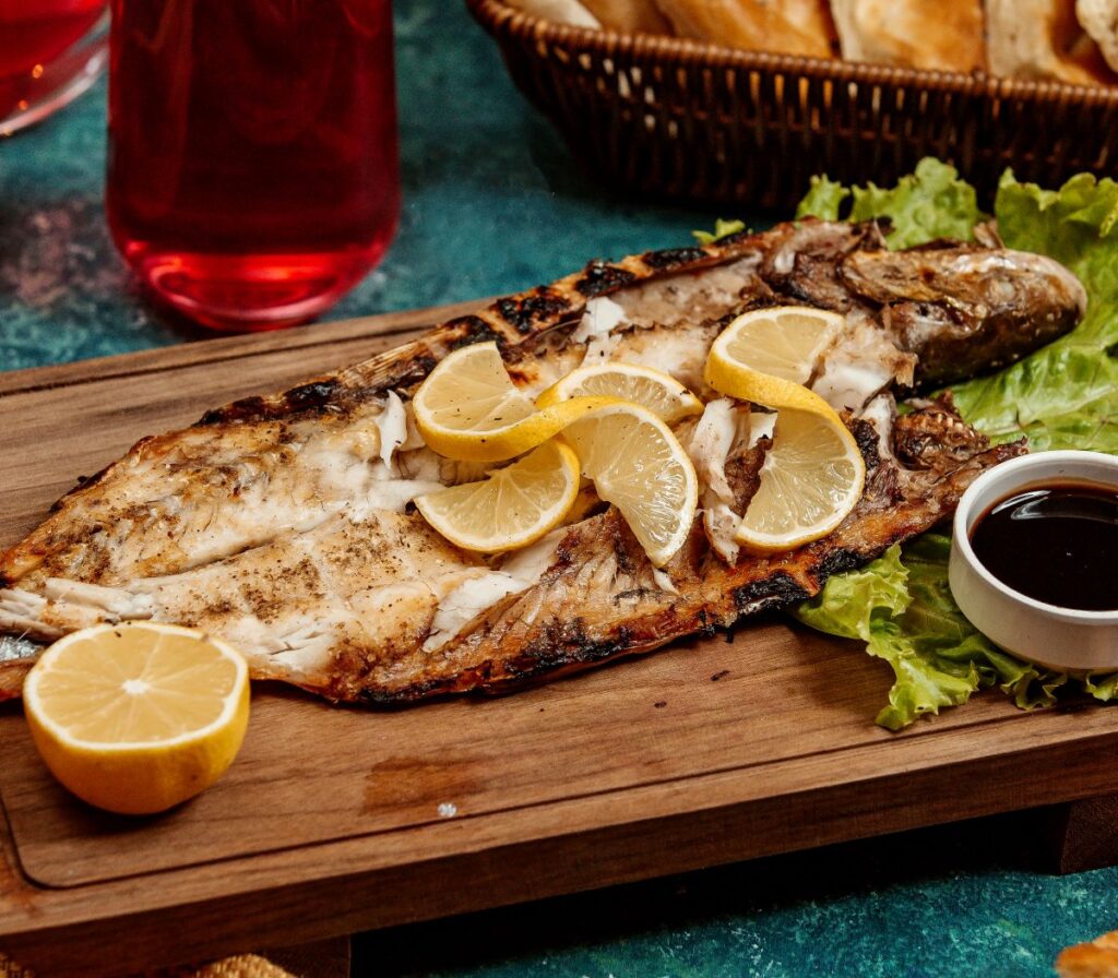 Quer variar o almoço? Aprenda receitas fáceis de preparar peixe assado no forno para o almoço 2 Peixe assado aberto com rodelas de limão sobre folhas de alface e molho ao lado.