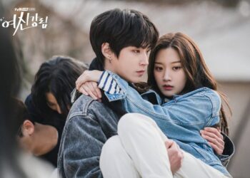 Jovem segura colega no colo com olhar protetor, ambos usam jaquetas jeans ao ar livre.