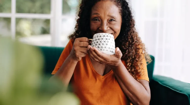 Café diário pode trazer benefícios para um envelhecimento mais saudável; entenda! 1 Mulher sênior sorri segurando xícara de café branca com estampa geométrica