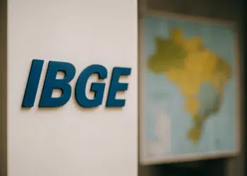 Letreiro azul do IBGE fixado em parede ao lado de mapa colorido do Brasil desfocado.