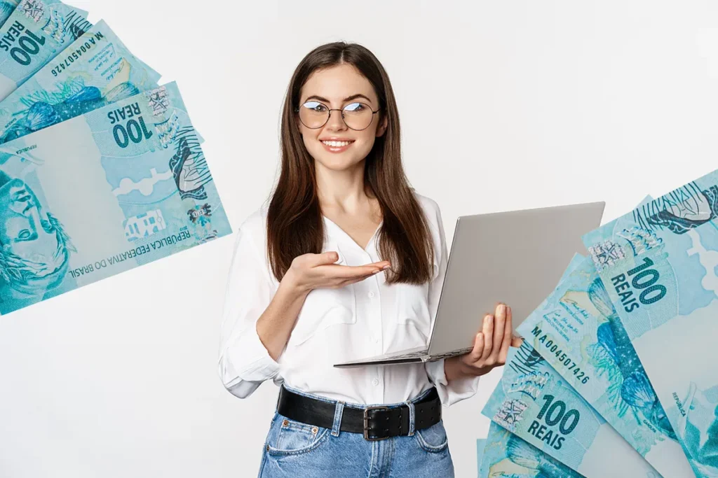 Valores esquecidos nos bancos somam R$ 10,5 bilhões; saiba como consultar e sacar seu dinheiro! 2 Mulher sorrindo segura notebook e aponta com mão, cédulas de R$100 ao redor.