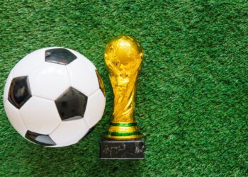 Bola de futebol posicionada ao lado de troféu dourado sobre gramado artificial.