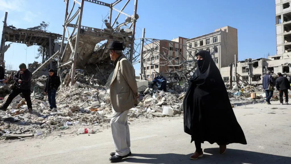 Guerra no Oriente Médio: ataques intensos marcam o 5º dia de conflito entre Irã, EUA e Israel 2 Pessoas atravessando rua em meio a prédios destruídos por ataques no conflito Irã, EUA e Israel