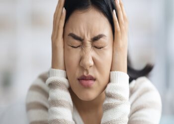 Mulher de olhos fechados pressiona as mãos nas orelhas e demonstra desconforto com o som de um apito.