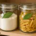 Dois potes de vidro com tampa, um contendo farinha e outro macarrão penne, ambos decorados com uma folha de louro sobre o vidro.