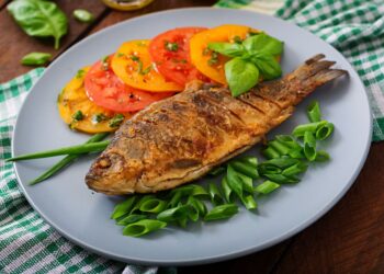 Peixe assado inteiro em prato cinza com tomates e ervas ao lado.
