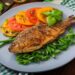 Peixe assado inteiro em prato cinza com tomates e ervas ao lado.