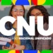 Duas jovens sorridentes, com óculos, seguram livros diante de painel colorido do CNU.