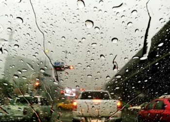 Visão através de um para-brisa coberto por gotas grossas de chuva, mostrando carros com luzes de freio acesas em uma via urbana congestionada durante o dia.