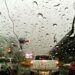 Visão através de um para-brisa coberto por gotas grossas de chuva, mostrando carros com luzes de freio acesas em uma via urbana congestionada durante o dia.