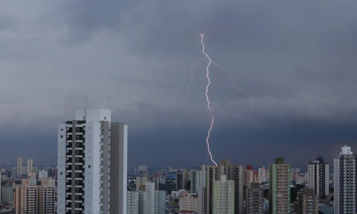 Alerta do Inmet: tempestades podem atingir diversas áreas do Brasil 2 Relâmpago vertical cruzando o céu cinzento e atingindo o topo de um edifício em uma área densamente urbana com vários prédios residenciais.