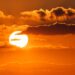 Fim de semana terá temporais, chuva forte e muito calor; confira a previsão 12 Close-up de um sol circular e branco-amarelado muito brilhante, posicionado à esquerda, iluminando nuvens densas e escuras de cor laranja-avermelhada. O fundo é um céu laranja vibrante com algumas camadas de nuvens mais finas na parte inferior.